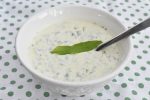 Creamy cucumber yogurt sauce with dill in a white bowl, topped with a green leaf and a spoon.