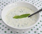 Creamy cucumber yogurt sauce with dill in a white bowl, topped with a green leaf and a spoon.