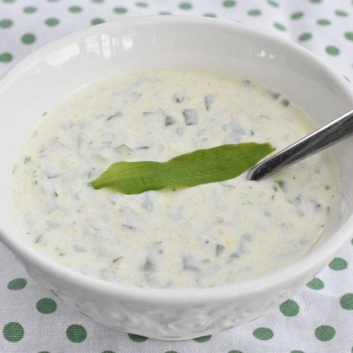 Creamy cucumber yogurt sauce with dill in a white bowl, topped with a green leaf and a spoon.