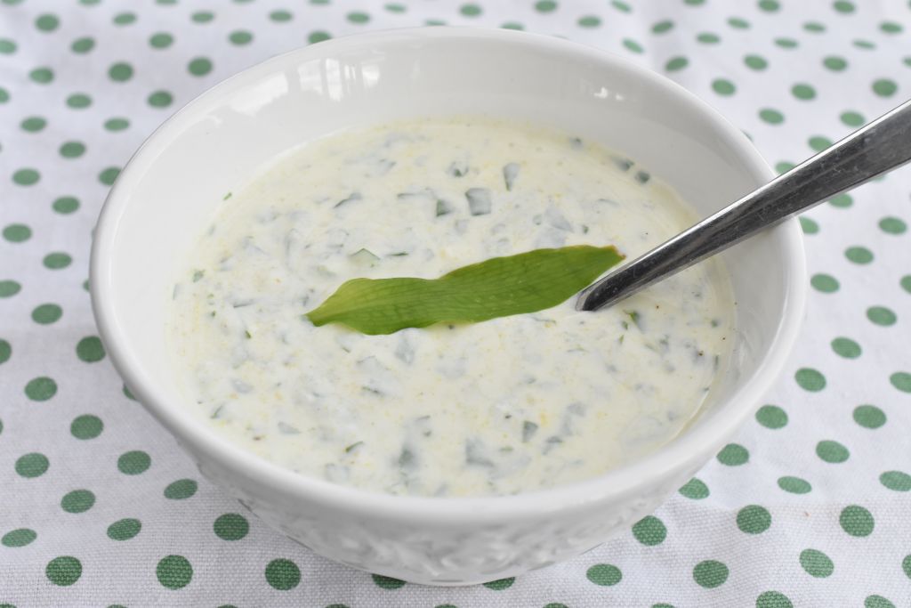 Creamy cucumber yogurt sauce with dill in a white bowl, topped with a green leaf and a spoon.